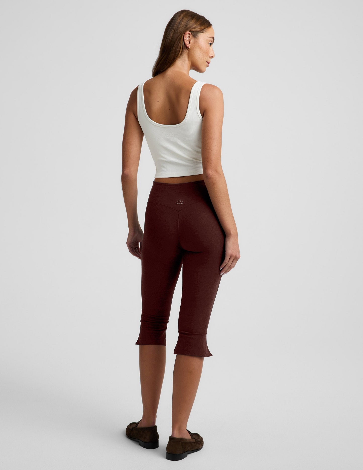 Woman wearing a white tank top and brown pedal pushers on a plain background.