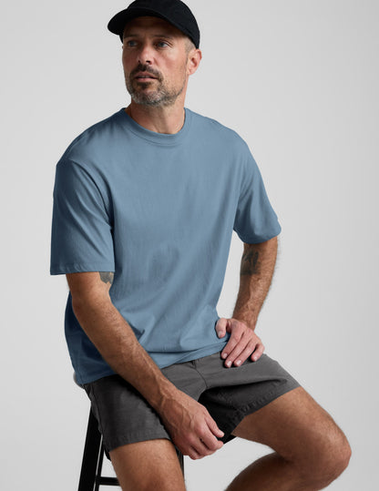 A man sitting on a stool wearing the gray Saturday Men's Short and a blue t-shirt. The shorts have a mid-thigh length.