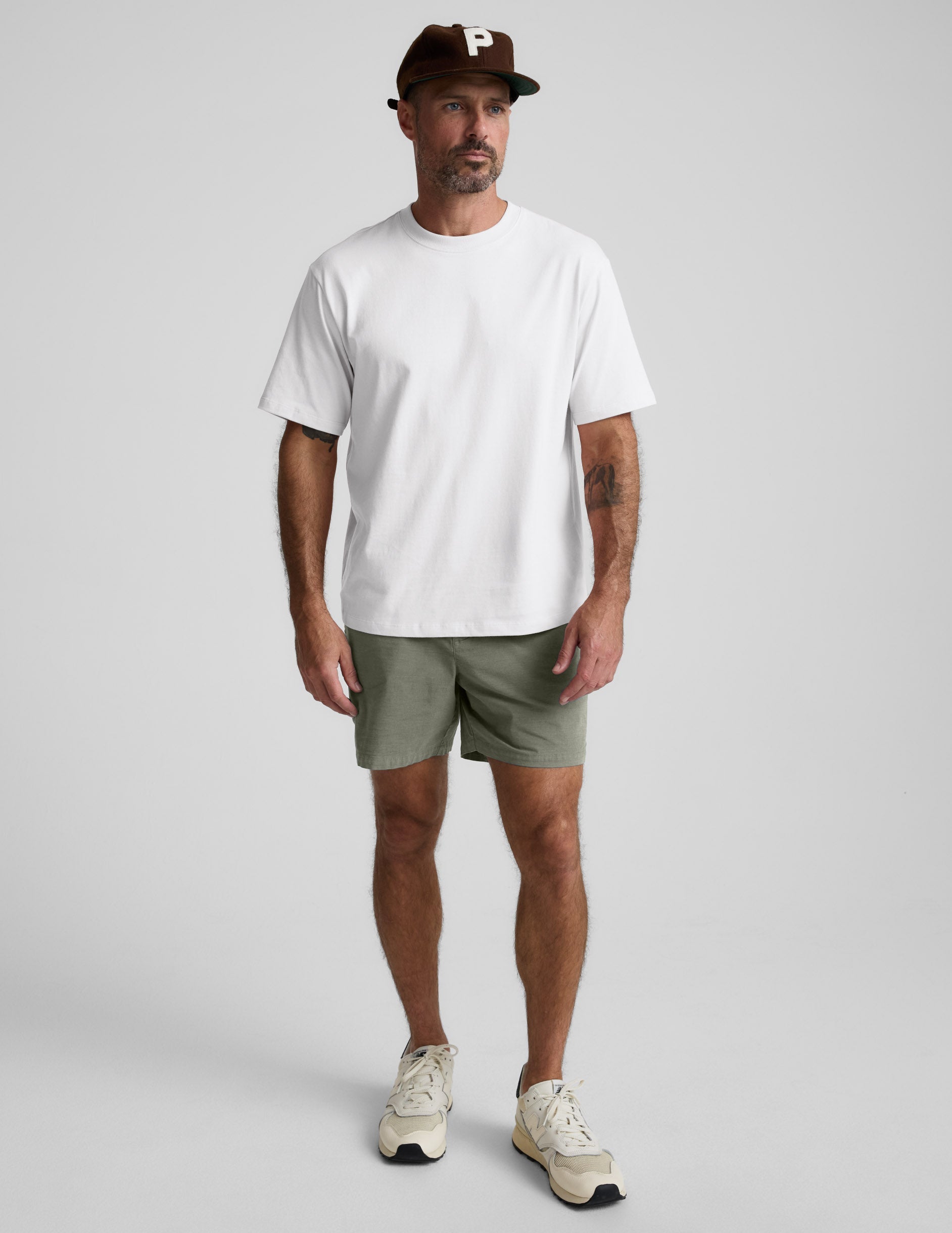 Man wearing olive Saturday Men's Short with a white t-shirt and brown cap, standing against a light grey background.