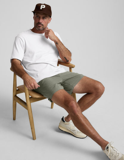Man sitting in a chair wearing olive green Saturday Men's Short, a white t-shirt, and a brown cap.