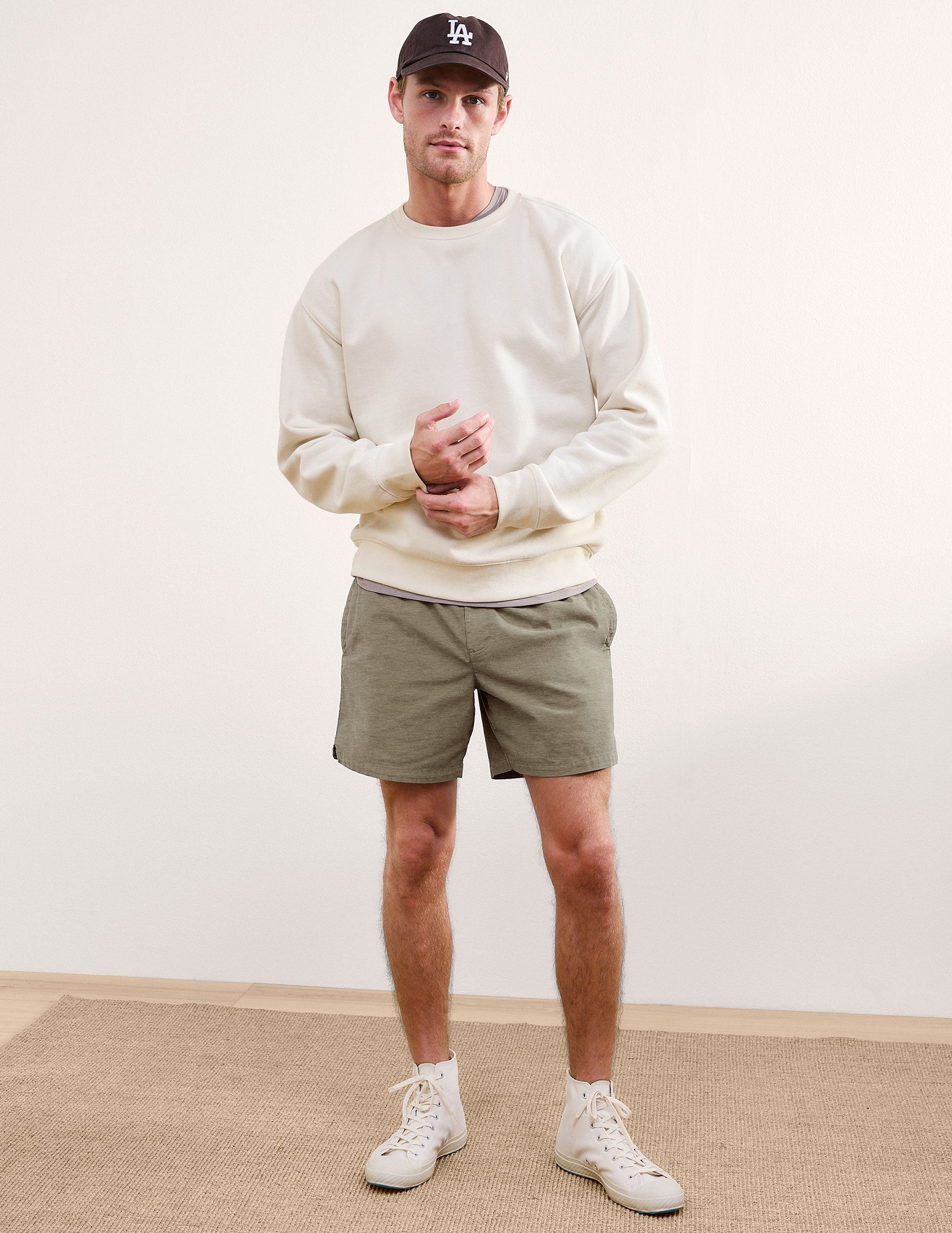 Front view of olive Saturday Men's Short on a model, paired with a cream sweatshirt, baseball cap, and white sneakers.