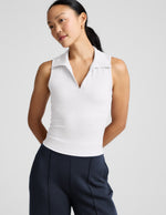 Woman wearing a white polo tank with a cutout in the back and blue pants against a plain background.