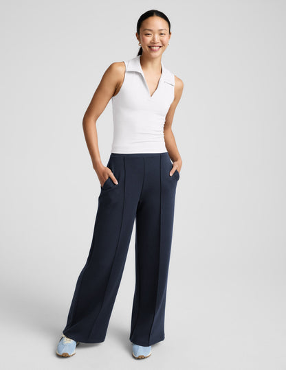 Woman wearing a white polo tank with a cutout in the back and blue pants against a plain background.