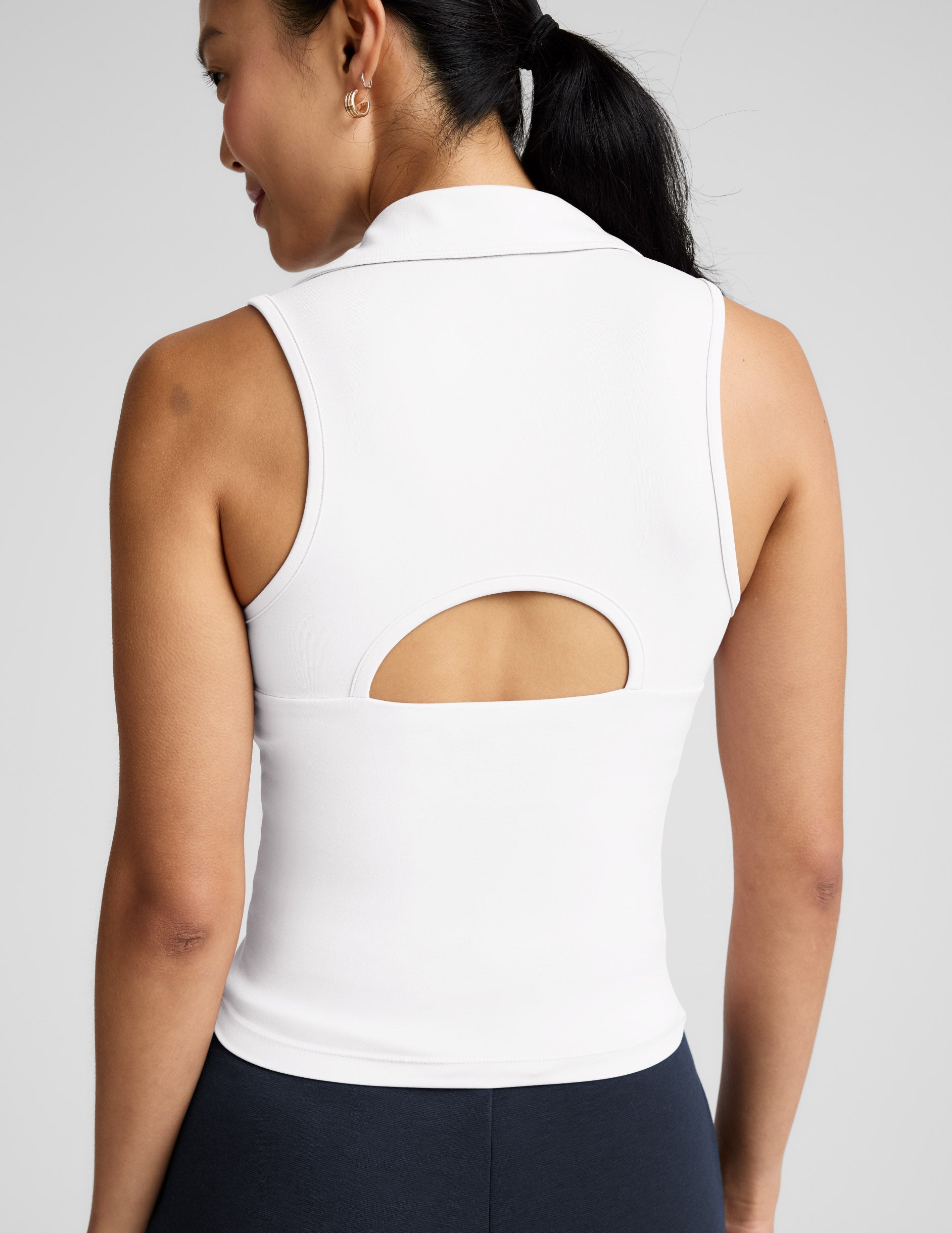 Woman wearing a white polo tank with a cutout in the back and blue pants against a plain background.