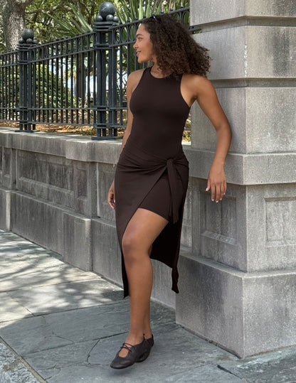 A woman wearing a dark brown sleeveless wrap-style dress with a high slit and side tie, standing outdoors against a gray stone wall and black metal fence.