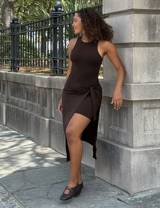 A woman wearing a dark brown sleeveless wrap-style dress with a high slit and side tie, standing outdoors against a gray stone wall and black metal fence.