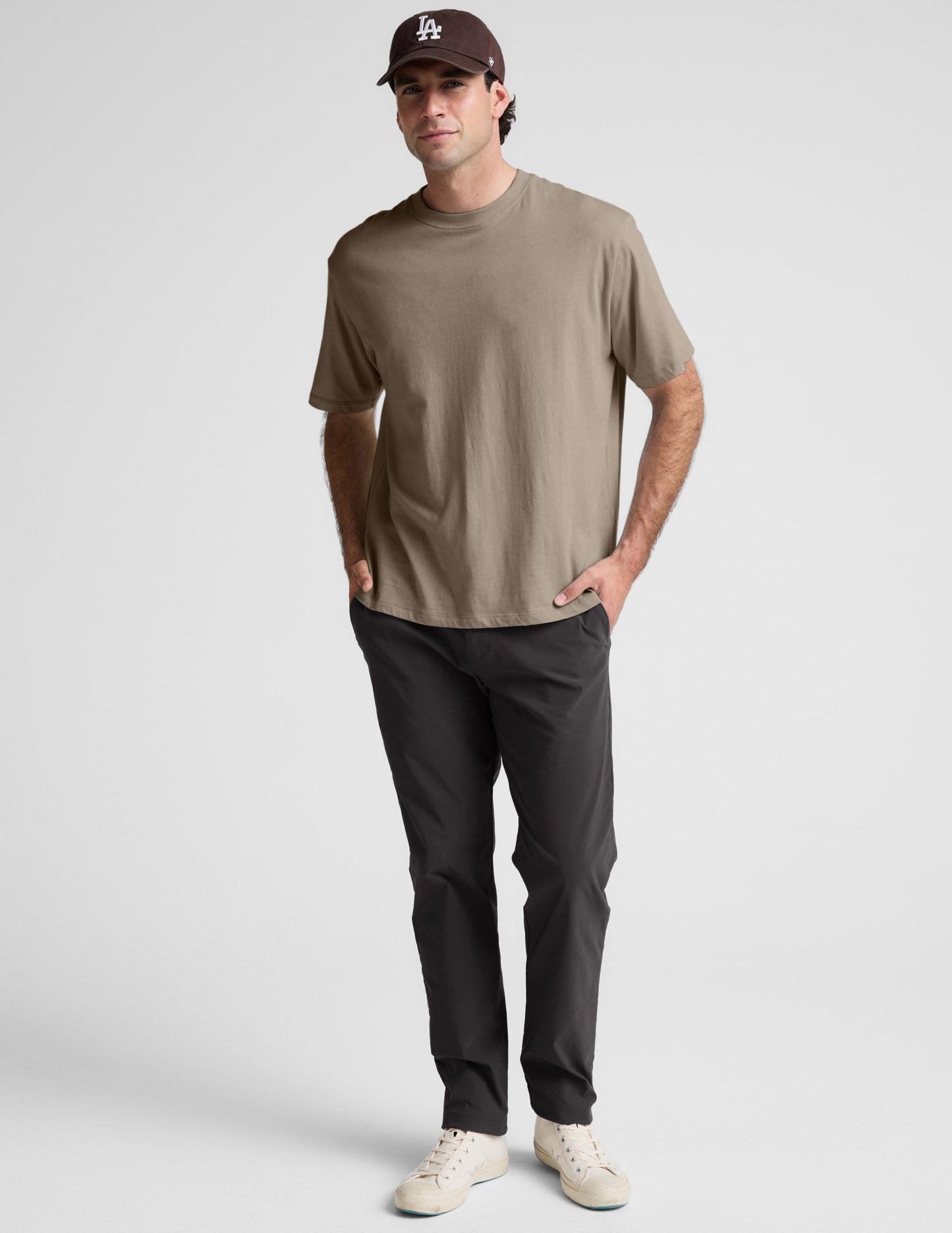 Man wearing a brown t-shirt, black pants, and a cap on a light gray background.