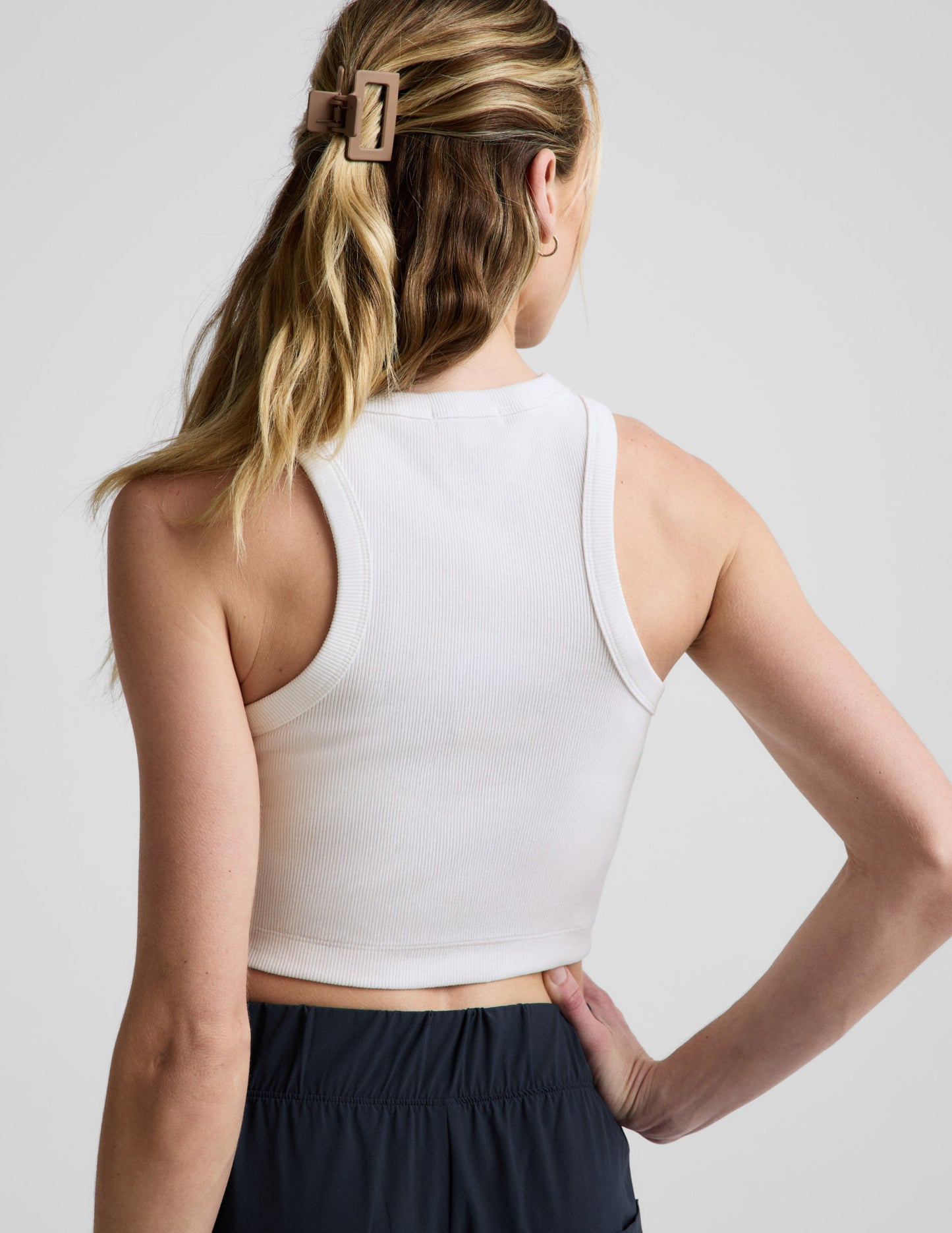 Woman wearing a white ribbed cropped tank and blue pants against a plain background.