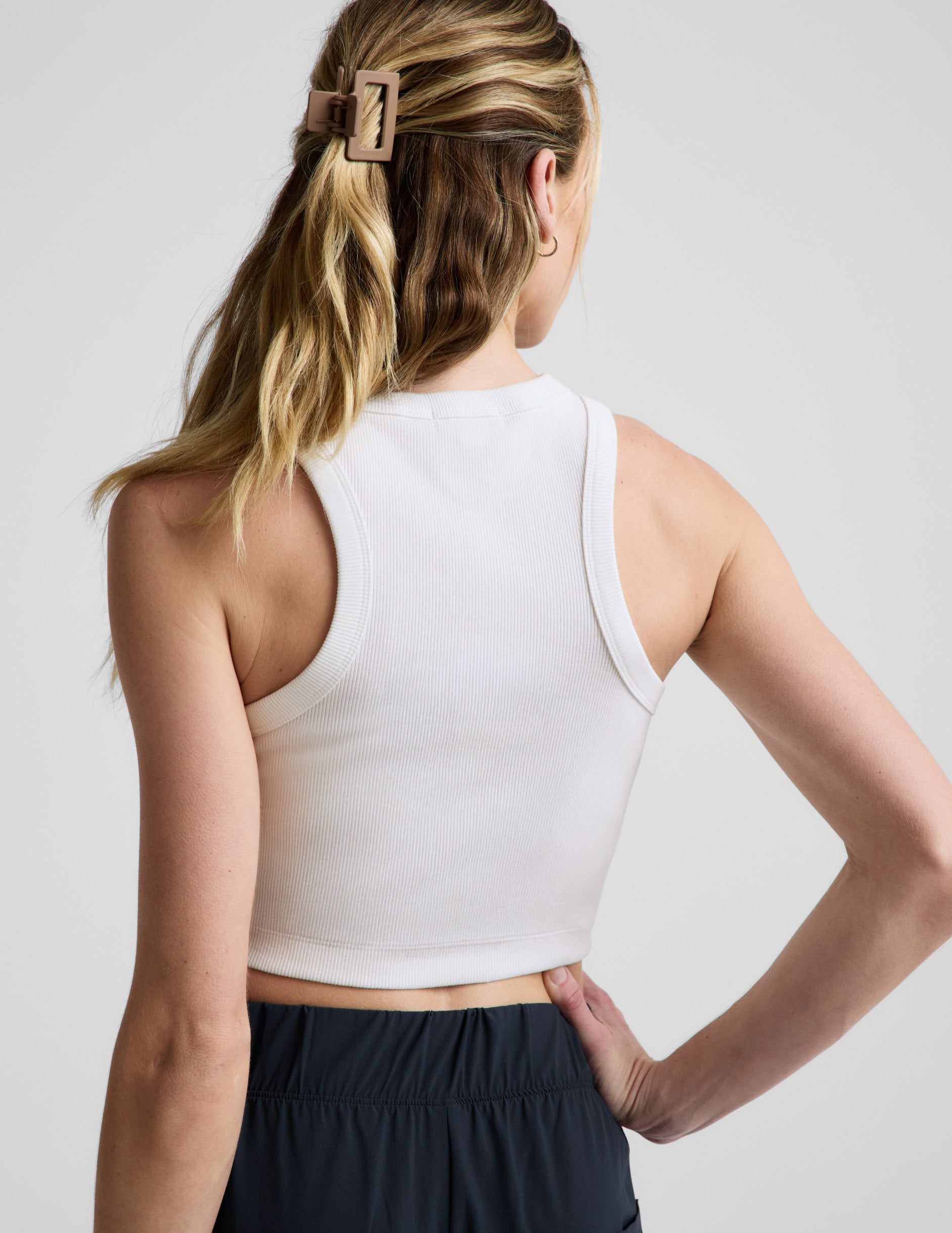 Woman wearing a white ribbed cropped tank and blue pants against a plain background.