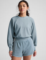 Woman wearing a blue crew-neck sweatshirt and shorts against a plain background.