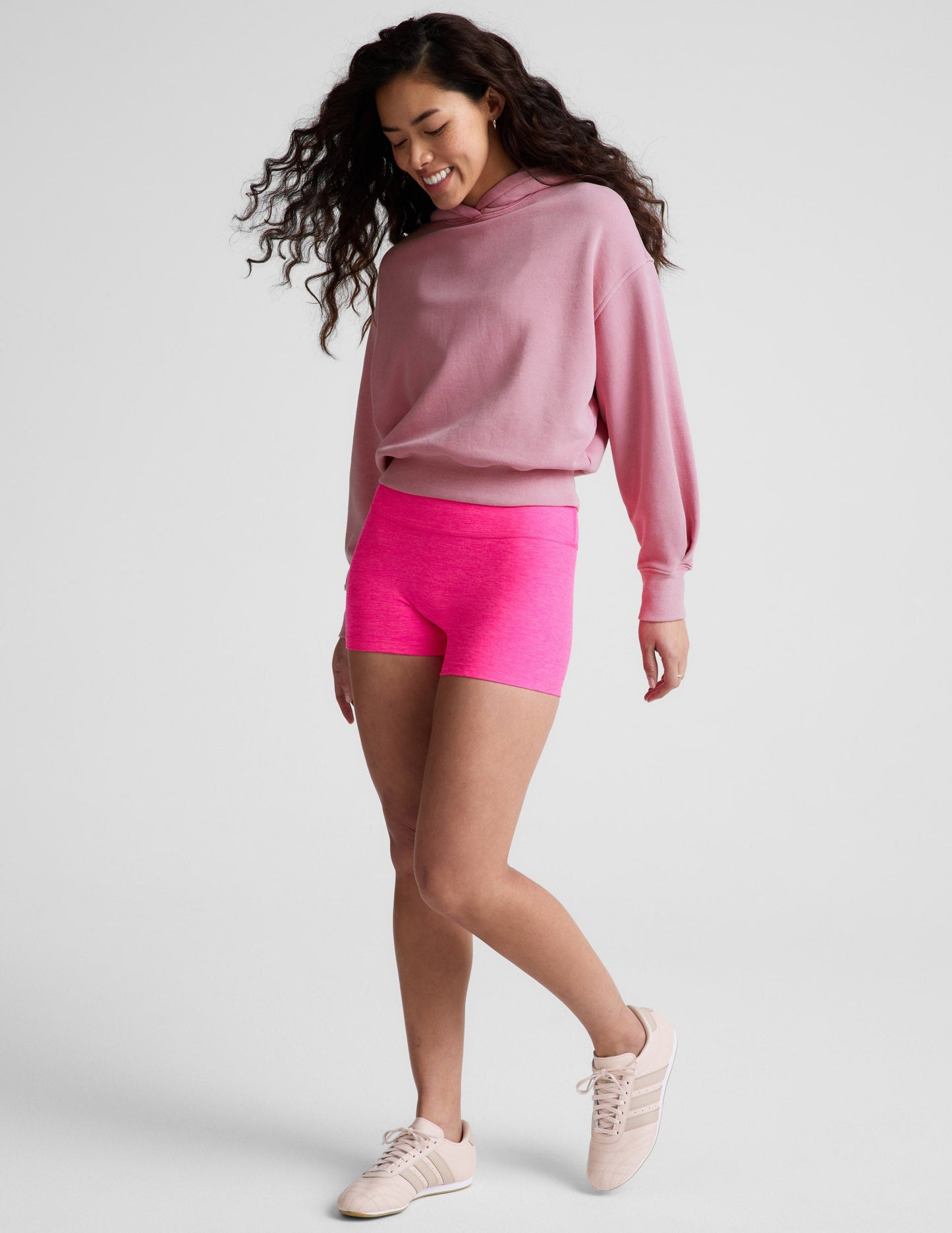 Woman wearing a pink sweatshirt and bright pink shorts on a plain background.