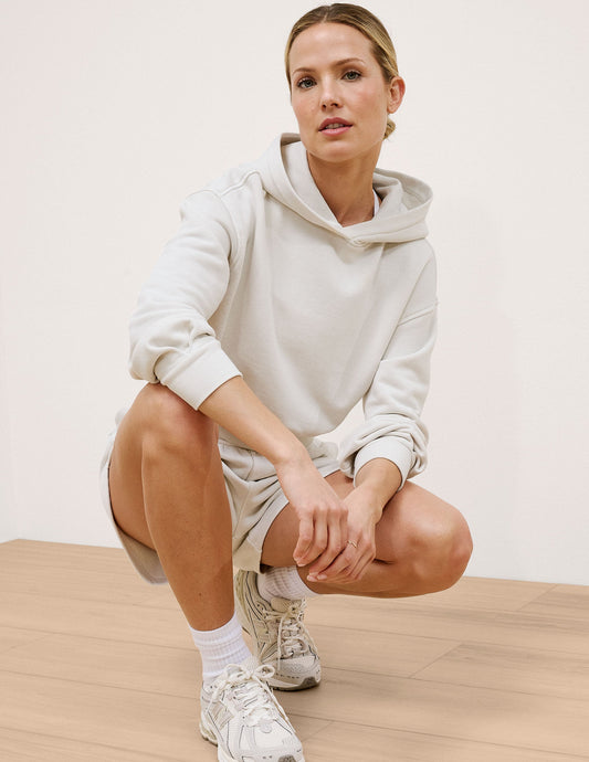 Woman wearing a white hoodie and shorts sitting on a wooden floor.