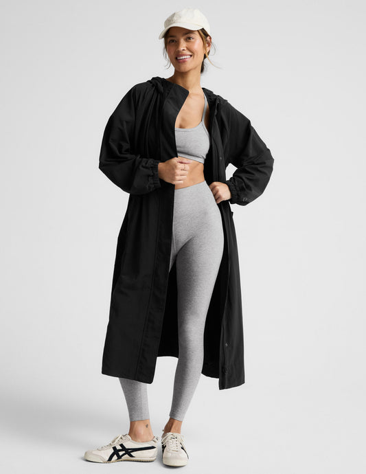 Woman wearing a black trench coat over a gray sports bra and leggings on a plain background.
