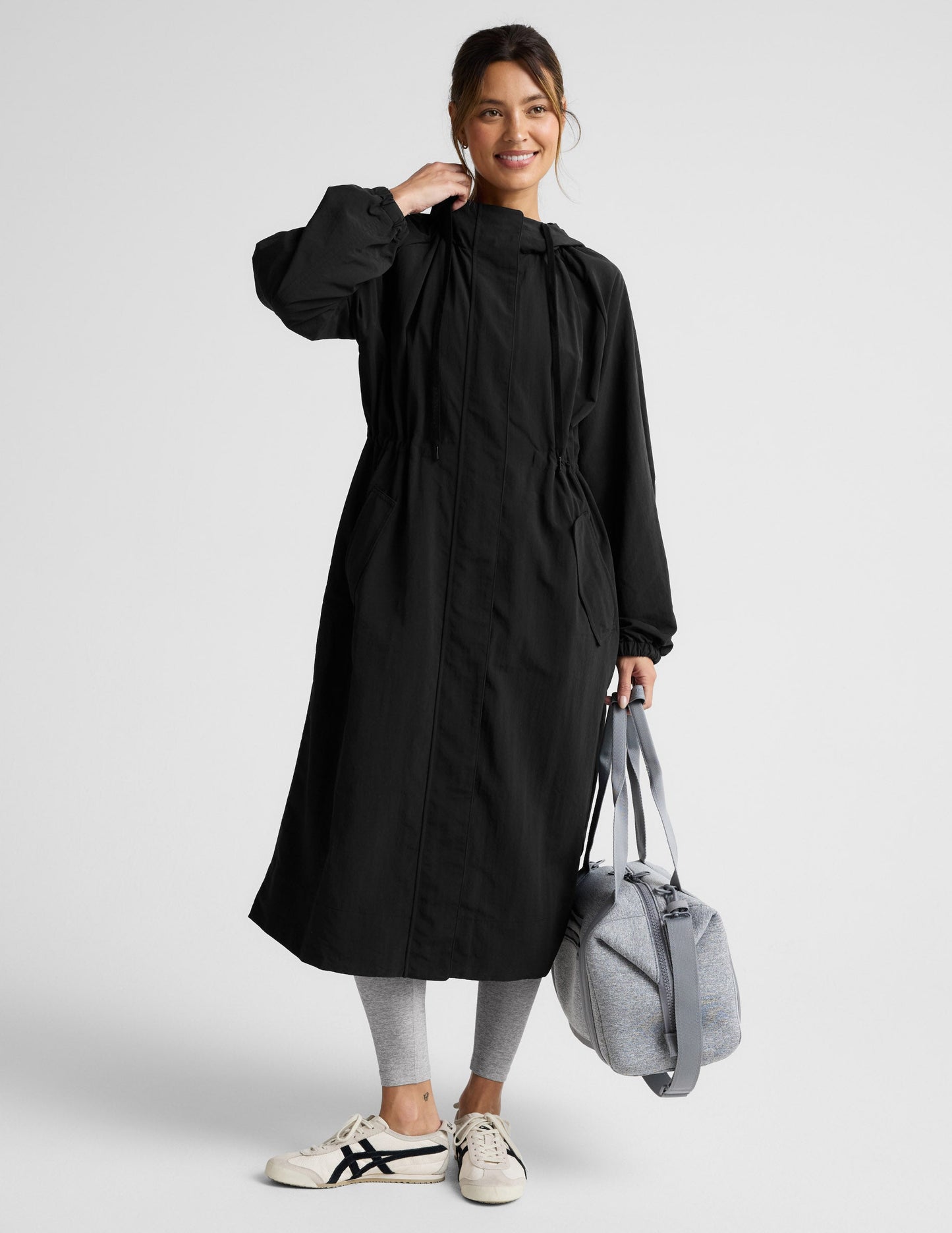 Woman wearing a black trench coat over a gray sports bra and leggings on a plain background.