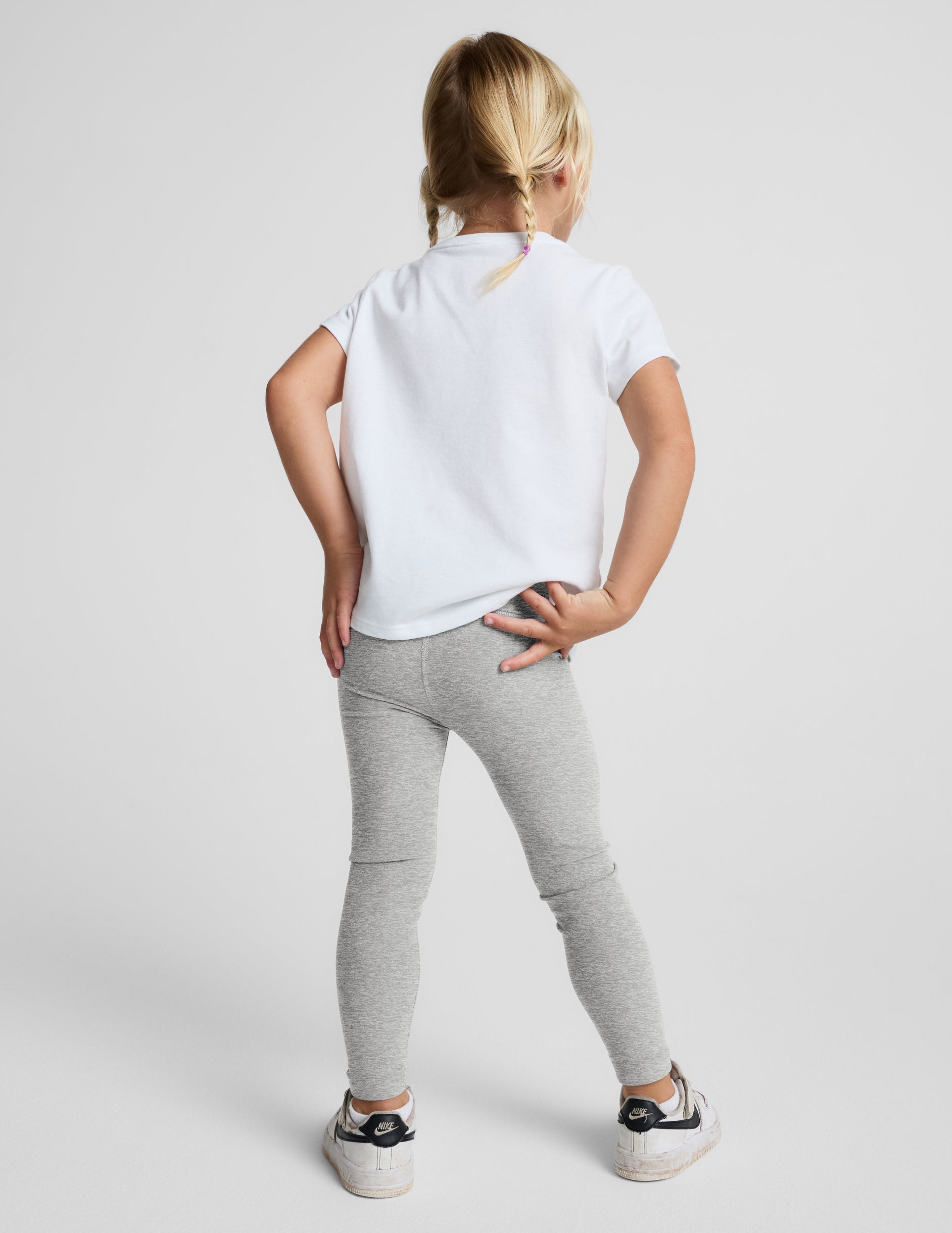 A young girl with blonde braids stands against a light gray studio background wearing a white short-sleeve t-shirt, light gray heathered leggings, and white sneakers.