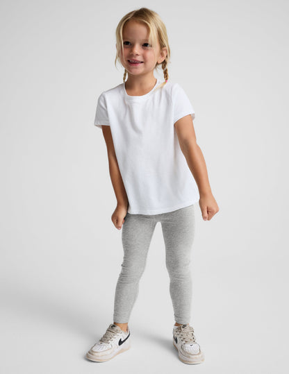 A young girl with blonde braids stands against a light gray studio background wearing a white short-sleeve t-shirt, light gray heathered leggings, and white sneakers.