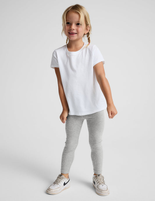 A young girl with blonde braids stands against a light gray studio background wearing a white short-sleeve t-shirt, light gray heathered leggings, and white sneakers.