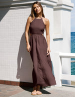 Woman in a brown linen halter dress standing against a white wall with ocean view.