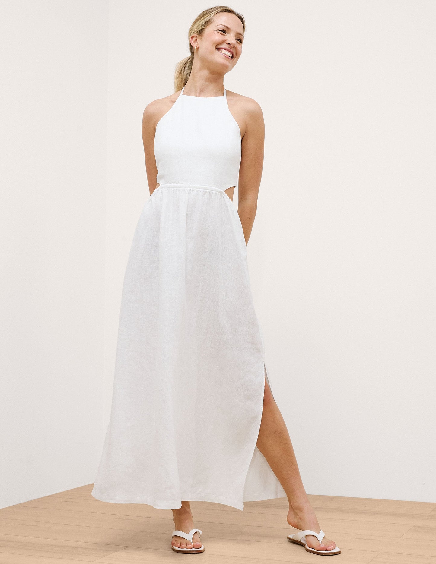 Woman wearing a white halter neck dress with a slit on a plain background.