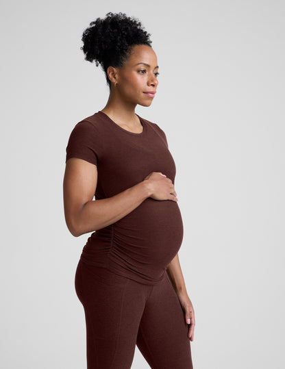 Pregnant woman wearing a brown maternity t-shirt and leggings against a plain background.