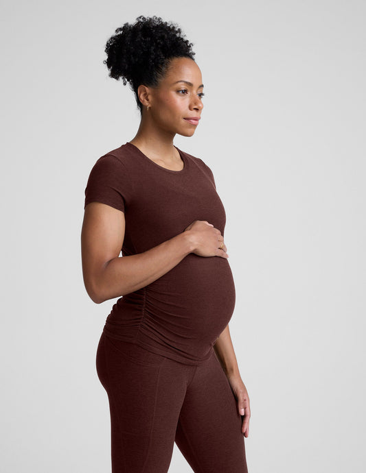 Pregnant woman wearing a brown maternity t-shirt and leggings against a plain background.