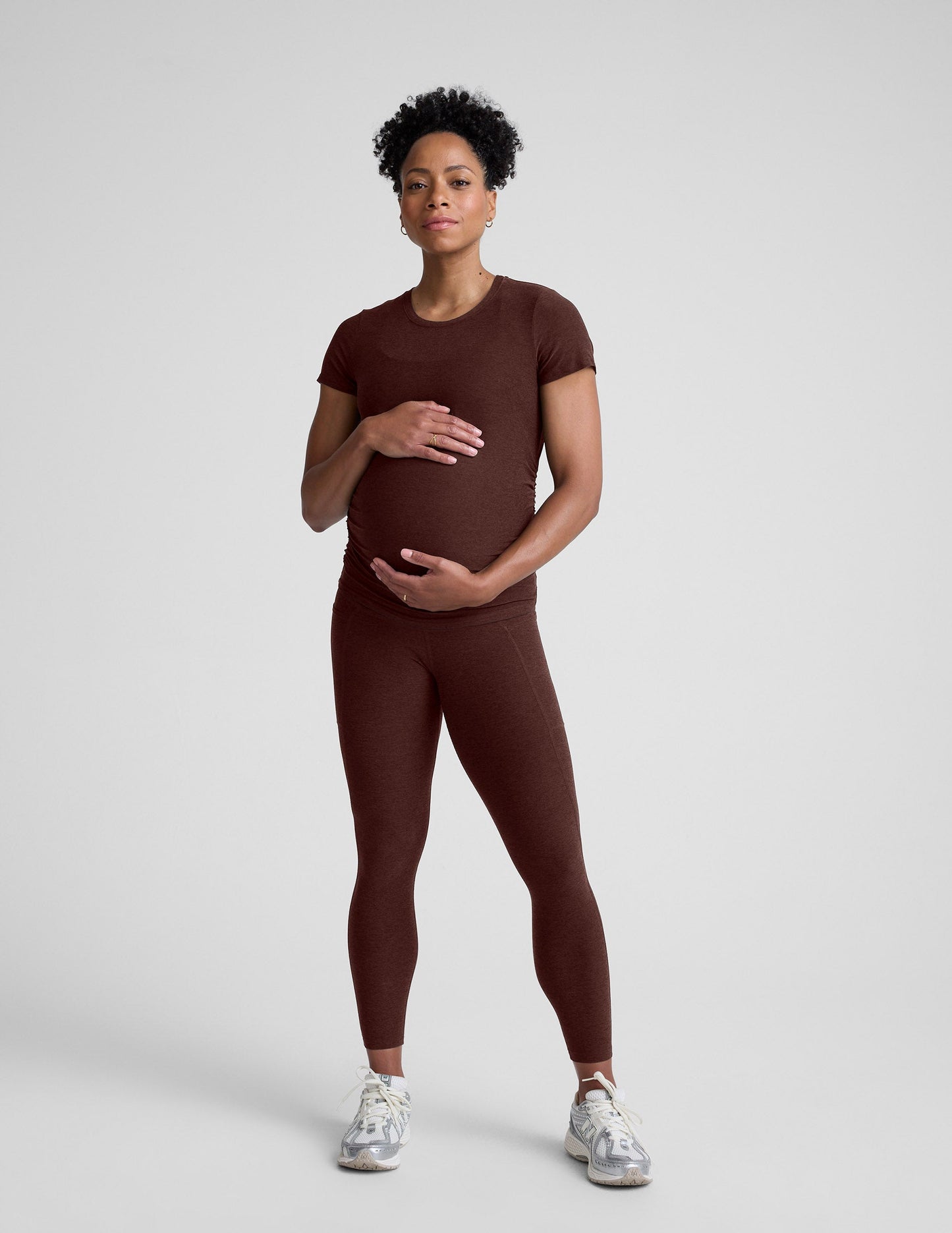 Pregnant woman wearing a brown maternity t-shirt and leggings against a plain background.