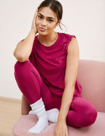 Woman in a magenta t-shirt and leggings outfit sitting on a pink couch with a white background.