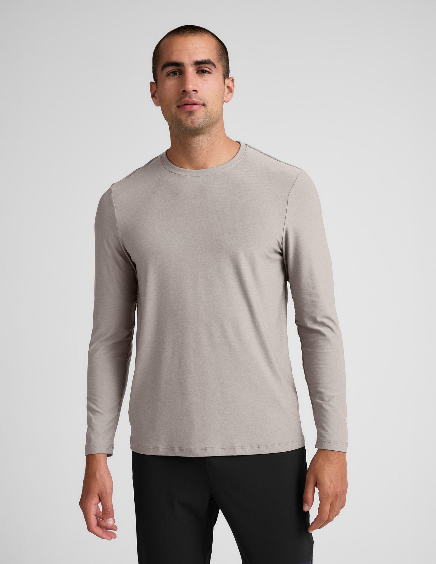 Man wearing a beige long-sleeve shirt and black pants against a plain background.