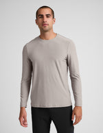 Man wearing a beige long-sleeve shirt and black pants against a plain background.