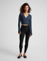 Woman wearing a blue long-sleeve wrap top and black leggings on a plain background.