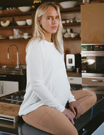 Woman sitting on a kitchen counter wearing a white maternity shirt and beige leggings.