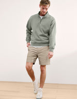 Man wearing beige Element Men's Chino Short 7" with a green quarter-zip and white sneakers, walking in a bright room.