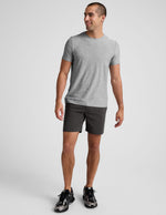Man wearing dark grey Essential Men���s Chino 7��� Short with a grey tee and sneakers, standing against a light grey backdrop.