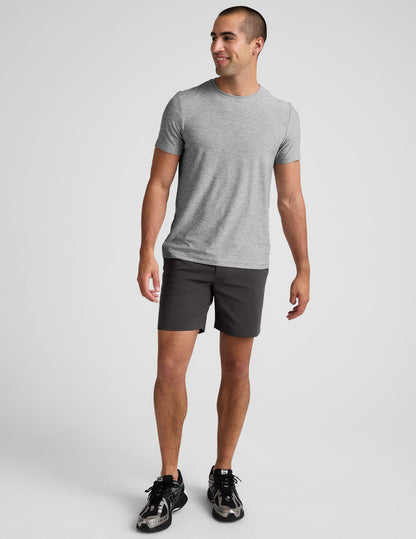 Man wearing dark grey Essential Men���s Chino 7��� Short with a grey tee and sneakers, standing against a light grey backdrop.