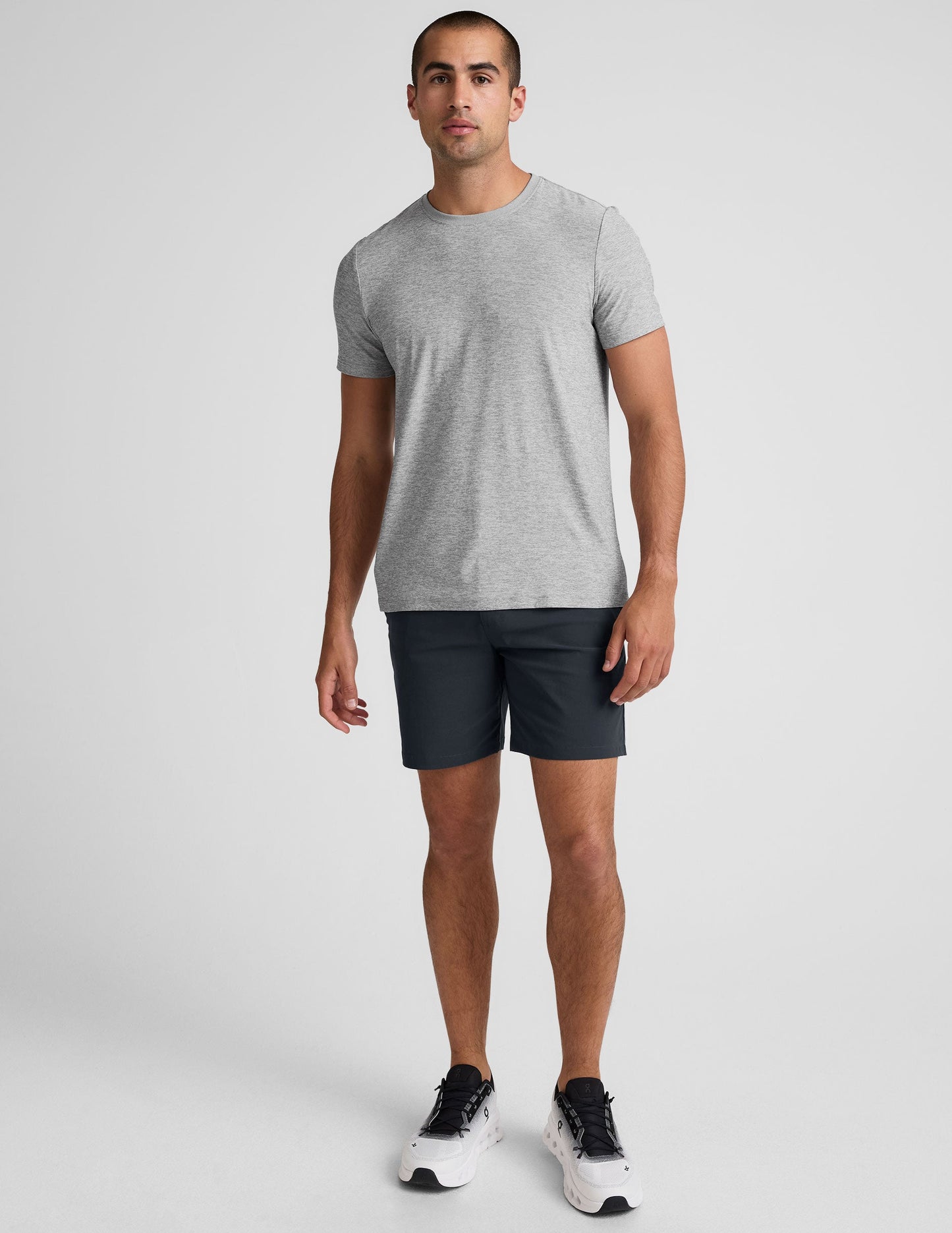 A man wearing dark grey Essential Men's Chino 7" Shorts, a grey t-shirt, and sneakers against a white background.
