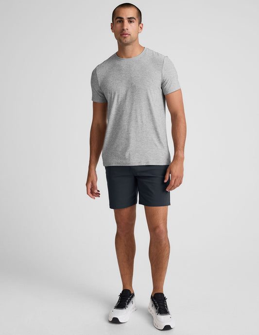 A man wearing dark grey Essential Men's Chino 7" Shorts, a grey t-shirt, and sneakers against a white background.