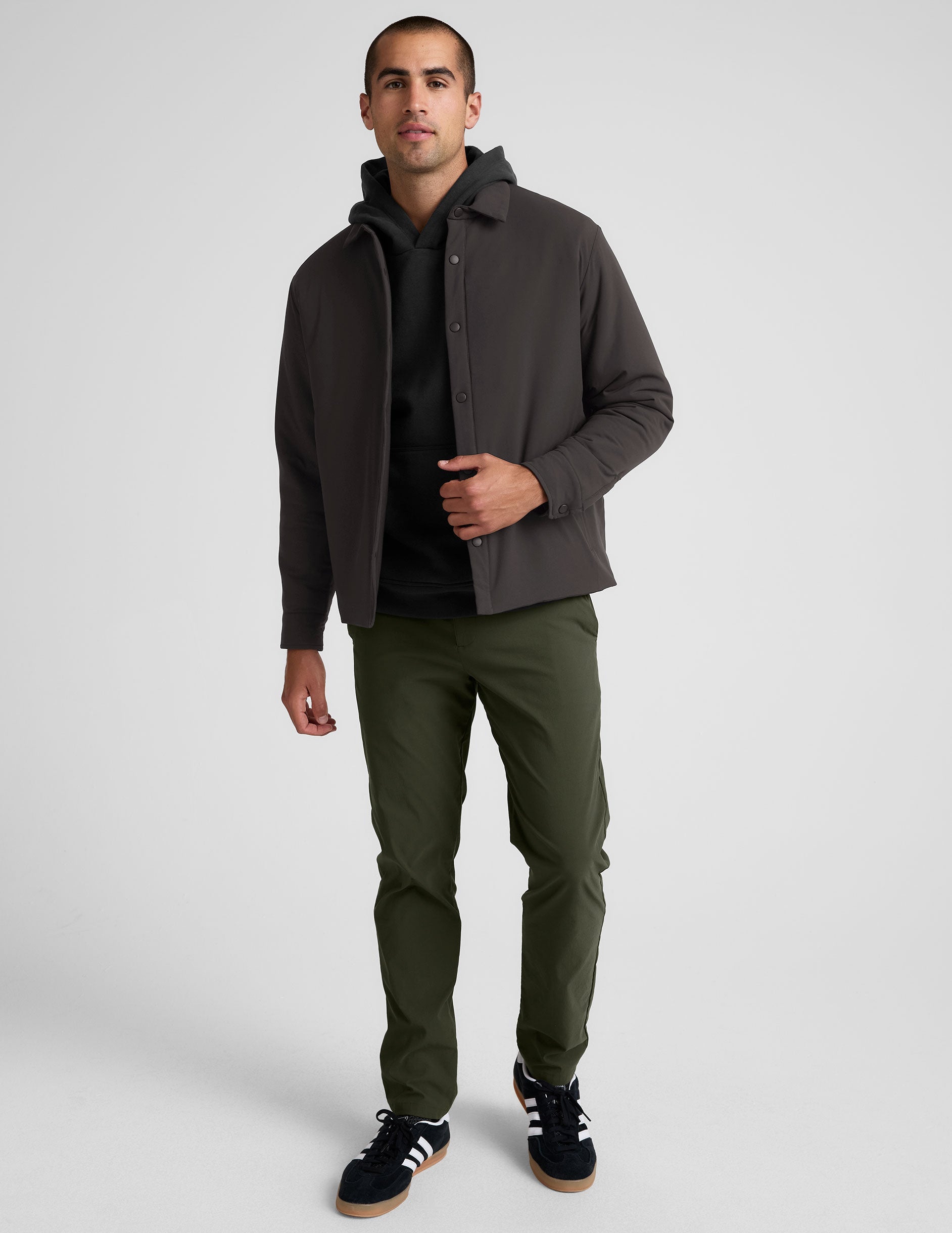 Man wearing a dark jacket, black shirt, green pants, and black sneakers on a white background.