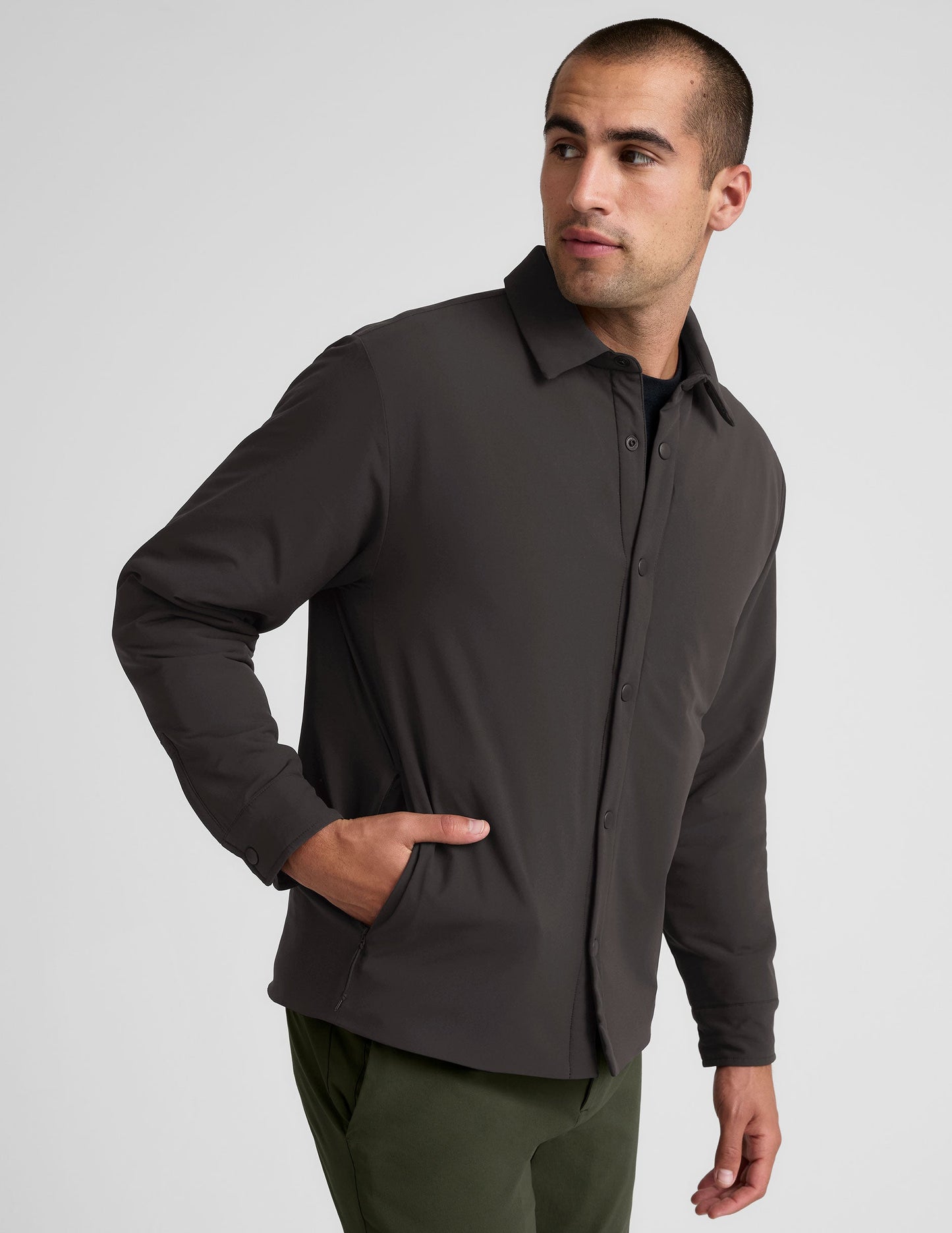 Man wearing a dark brown jacket over a black shirt with green pants on a light gray background.