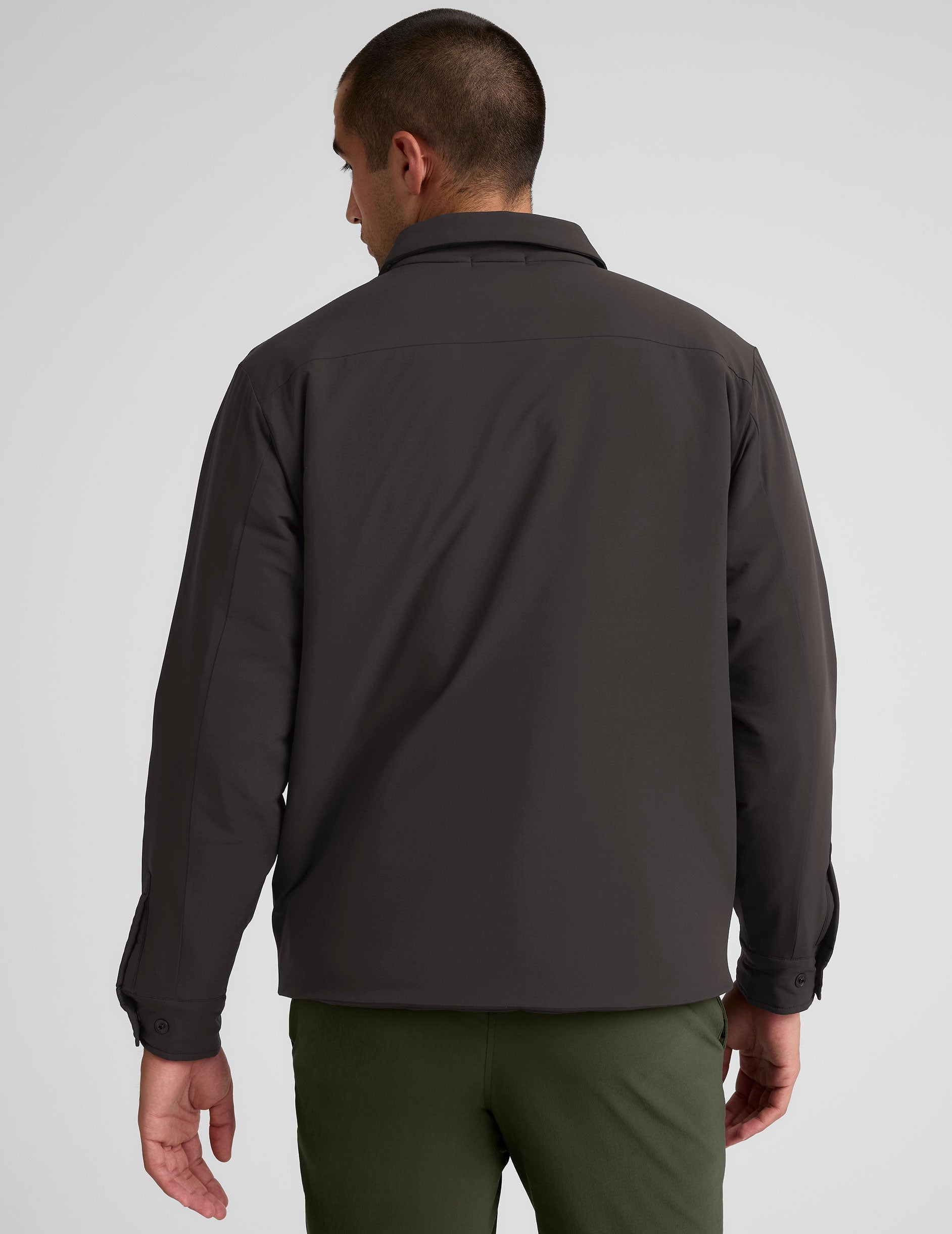 Man wearing a dark brown jacket over a black shirt with green pants on a light gray background.