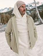 Man wearing a beige jacket and white hoodie in a snowy landscape with mountains.