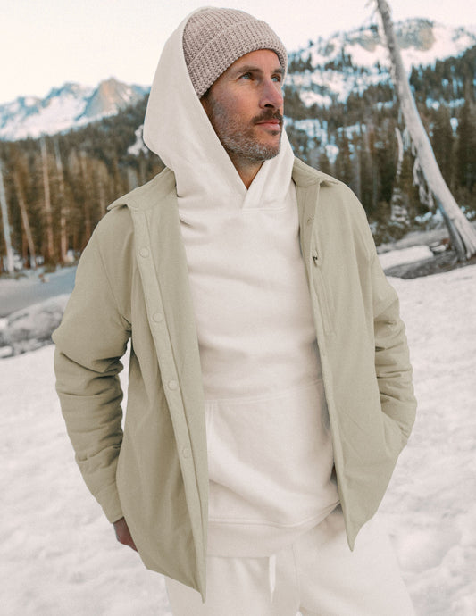 Man wearing a beige jacket and white hoodie in a snowy landscape with mountains.