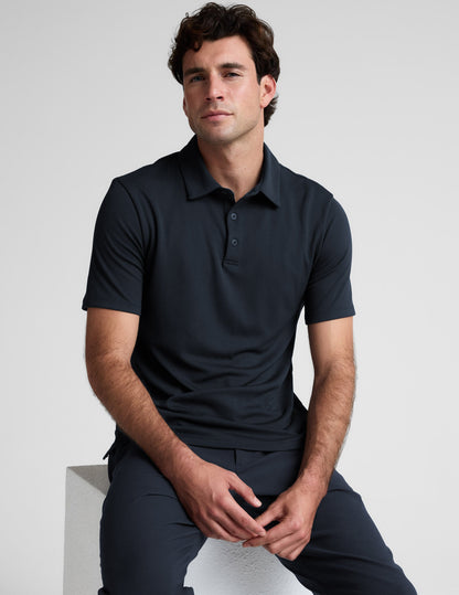 Man wearing a dark blue polo shirt and pants sitting on a white block against a light gray background.