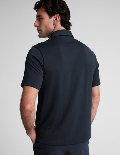 Man wearing a dark blue polo shirt and pants against a light gray background.