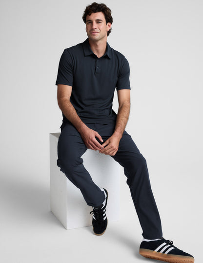 Man wearing a dark blue polo shirt and pants against a light gray background.
