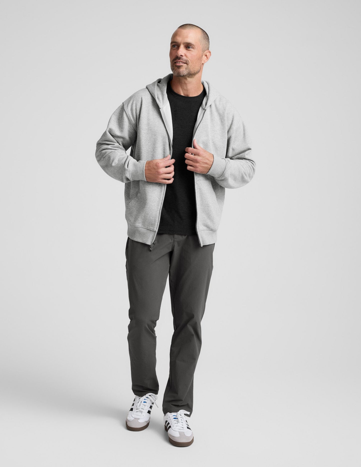 Man wearing a gray full zip hoodie and dark gray pants on a plain background.