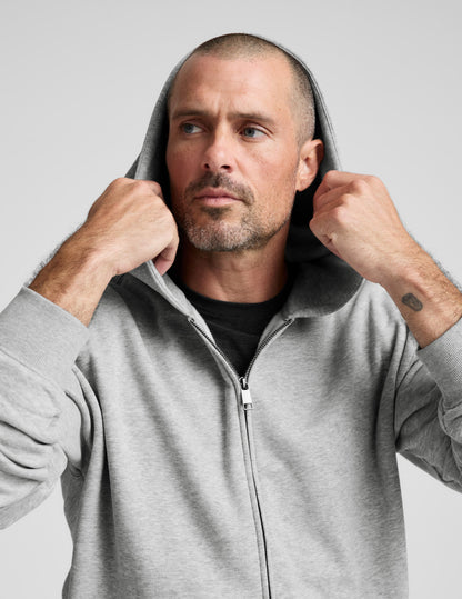 Man wearing a gray full zip hoodie and dark gray pants on a plain background.