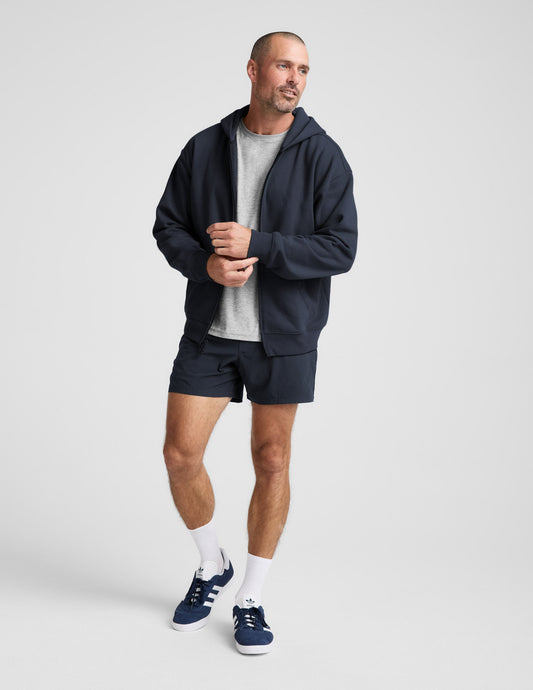 Man wearing a navy blue full zip hoodie and shorts on a plain background.