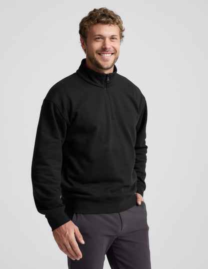 Man wearing a black quarter-zip sweatshirt and gray pants on a light gray background.