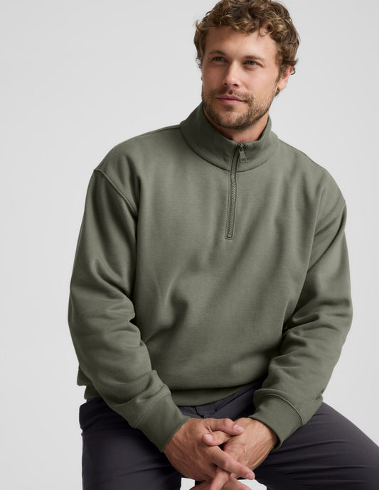 Man wearing a green quarter zip pullover and khaki pants with a plain background.