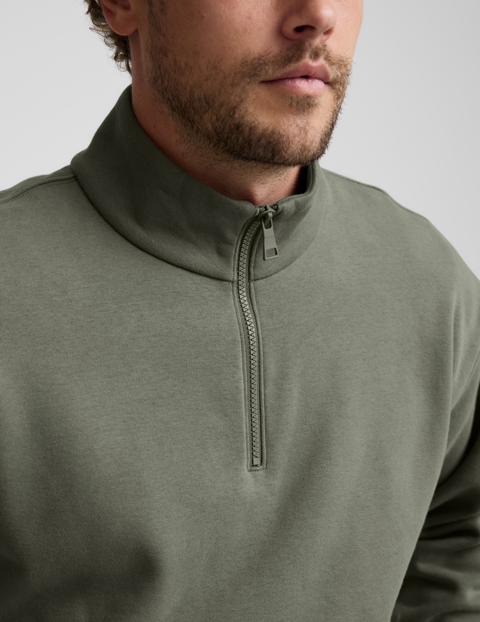 Man wearing a green quarter zip pullover and khaki pants with a plain background.